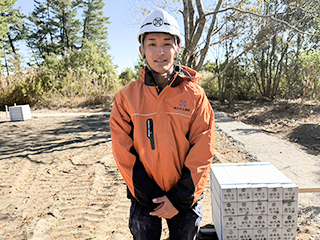 オレンジの作業服と白いヘルメットを着用し、建設現場で笑顔を浮かべる現場監督