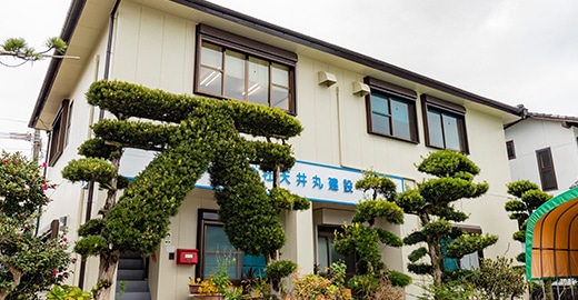 株式会社天井丸建設のオフィス外観。クリーム色の2階建ての建物に青い看板、手入れの行き届いた庭