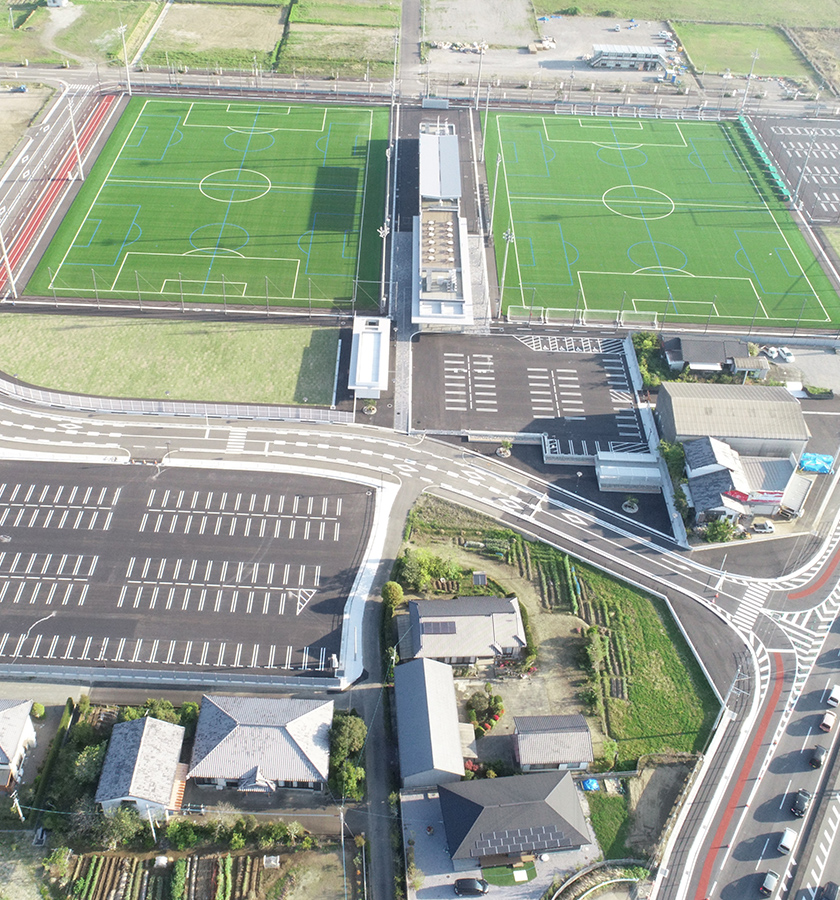 緑の人工芝サッカーグラウンド2面と中央のクラブハウス、広い駐車場が整備されたスポーツ施設の空撮写真