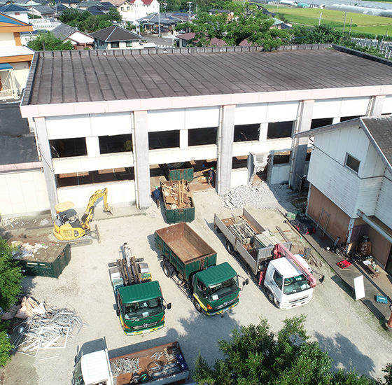 大型の白い建物を解体する解体工事の現場。黄色いショベルやダンプトラック、廃材コンテナが配置された空撮写真