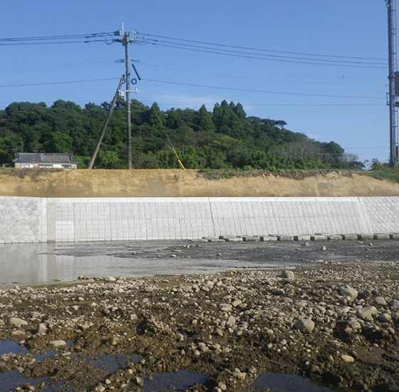 コンクリートブロックによる護岸整備が施された河川工事の施工例。淡い灰色のブロック壁と緑の丘陵が映る