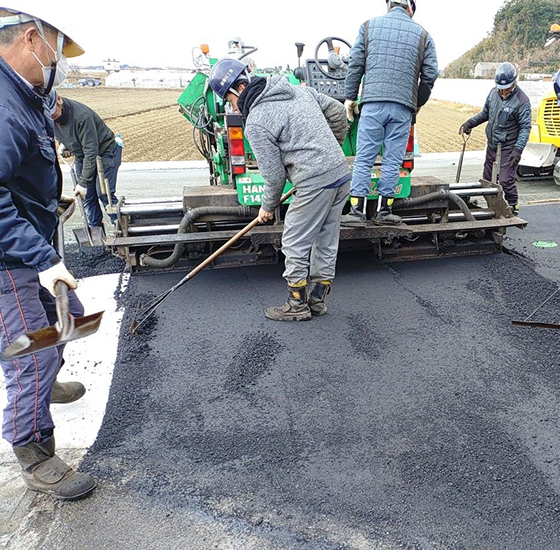アスファルト敷設機で道路を舗装する作業員たち。ショベルやレーキで新たに敷いたアスファルトを均す道路工事の様子